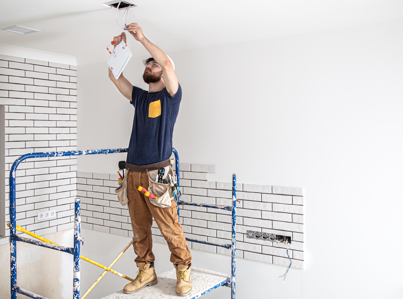 Elektriker Builder bei der Arbeit, Installation von Lampen in der Höhe. Fachmann im Overall mit einem Elektrowerkzeug. Im Hintergrund der Reparaturstelle. Das Konzept der Arbeit als Profi.